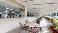 This single-story facility is designed to serve as a communal amenity space, combining indoor and outdoor areas in a tropical setting. The covered outdoor patio runs along the length of the building, featuring a broad, white paneled ceiling that provides shade while maintaining an open-air feel. Multiple round tables with slatted metal chairs are arranged along the patio, creating a comfortable seating area ideal for socializing or relaxing outdoors. Large glass windows and panels line the interior wall of the patio, offering clear sightlines into the fitness center within.

The fitness center is visible through the floor-to-ceiling glass partition, and is well-lit by overhead fluorescent lighting. It appears to house a variety of cardio and strength training equipment, making it a functional space for exercise. A glass entry door with a welcome mat provides direct access from the patio into the fitness area.

Beyond the patio, lush tropical landscaping frames the space, with vibrant shrubs, decorative red mulch beds bordered by natural stone, and tall palm trees creating a verdant backdrop. A community pool is visible in the near distance, enclosed by a white fence. The overall ambiance is bright, clean, and inviting, reflecting a well-maintained Florida-style community environment.