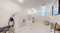 Located at 2226 North Davidson Street in Charlotte's NoDa neighborhood, this purpose-built pet grooming facility offers a clean, functional, and thoughtfully designed space for pet care. The single-room layout is finished with neutral cream-toned subway tile wainscoting that lines the walls, complemented by speckled terrazzo-style flooring that is both durable and easy to maintain. Bright fluorescent lighting illuminates the entire space, while a window along one wall allows natural light to filter in, adding an airy quality to the room. The wash area features a walk-in dog bathing station with tiled walls, a built-in hose and nozzle system, and floor drains for efficient water management. Adjustable hydraulic grooming tables are positioned throughout the room, providing practical workstations for professional grooming services. A sleek black metal shelving unit holds neatly rolled towels and an assortment of grooming products, keeping essentials organized and accessible. Framed portrait artwork of various dog breeds adorns the upper walls, adding a welcoming and pet-friendly character to the space. The overall ambiance is clean, professional, and inviting, making this facility well-suited for both self-service and full-service pet grooming operations.