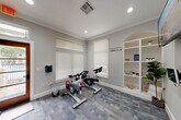 Located at 1895 Barker Cypress Rd in Houston, TX, this dedicated fitness room offers a well-equipped workout space designed to support a variety of exercise routines. The room is finished with cool gray walls, white crown molding, and a patterned gray tile floor that gives the space a clean, modern aesthetic. Recessed ceiling lights provide bright, even illumination throughout, complemented by natural light streaming in through multiple windows fitted with white horizontal blinds.

The space is equipped with two LynxCycle spin bikes positioned for group cycling sessions, alongside a multi-functional rack holding stacked stability balls in red, blue, and green, resistance bands, TRX straps, and an assortment of medicine balls. A weight bench adds further versatility for strength and conditioning workouts. A motivational wall decal reading "Don't wish for it. Work for it!" reinforces the room's energetic atmosphere.

A built-in arched shelving unit along one wall provides organized display storage, decorated with plants, informational materials, and a 24-hour video surveillance notice. A glass-paneled wooden door opens to an outdoor area with views of a pool and surrounding landscape, adding a refreshing connection to the outdoors. The space is under continuous video surveillance for added security.