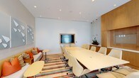 This single-floor space is designed as a contemporary conference and meeting room, combining functionality with a polished, professional aesthetic. At its center sits a generously sized rectangular conference table surrounded by ergonomic cream-upholstered chairs on casters, accommodating a sizeable group comfortably. A wall-mounted flat-screen display, framed in light wood paneling, serves as the focal point for presentations and collaborative work.

The room's clean white walls and recessed ceiling lighting create a bright, well-illuminated environment. Warm wood cabinetry lines one wall, providing storage and adding a refined, natural contrast to the neutral palette. A built-in credenza topped with a decorative plant introduces a touch of greenery, softening the professional setting.

A lounge area along one side features a sofa with orange accent cushions and a small side table, offering a more informal seating option. The floor is finished with a bold striped area rug in vibrant tones of orange, teal, and cream, injecting energy and visual interest into the space. Overall, the room balances a clean, modern design with warm, inviting details suited for focused meetings and presentations.