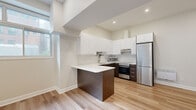 Located at 3474 Rue Hutchison in the heart of Montreal's Mile End neighbourhood, this single-floor unit offers a thoughtfully designed modern interior. The open-concept kitchen is a standout feature, showcasing a striking two-tone design with sleek white upper cabinetry and rich dark wood lower cabinets and island. A white countertop peninsula provides additional workspace and a natural gathering point. Stainless steel appliances, including a full-size refrigerator, stove, and built-in microwave, complete the contemporary kitchen setup. A tiled backsplash adds a refined finishing touch. Light hardwood flooring runs throughout the space, lending warmth and continuity across each area. Recessed lighting and large windows allow natural light to fill the interior, creating a bright and airy atmosphere. The living area is open and versatile, with clean neutral walls that offer a blank canvas. A well-proportioned hallway leads to additional rooms, each accessed through classic panelled white doors. The bathroom features full-height white tile, a soaking tub, and a multi-function brushed bronze shower panel system, combining functionality with a modern aesthetic. Overall, the space presents a clean, contemporary design well-suited for comfortable urban living in Montreal.