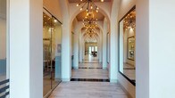 This thoughtfully designed single-floor space makes an immediate impression with its grand, arched hallway that serves as the architectural centerpiece. A series of graceful archways lines the corridor, creating a rhythmic, tunnel-like perspective that draws the eye toward a bright, open common area at the far end. Overhead, a procession of dramatic Sputnik-style chandeliers with warm Edison bulbs cast an inviting, ambient glow throughout the passage, adding a contemporary edge to the classical arch motif. The flooring features large-format light-toned tiles accented with darker inlay strips, lending the space a refined, polished character. To one side, floor-to-ceiling glass walls enclose office or meeting areas, maintaining visual openness while providing functional separation. Signage is visible through the glass, suggesting a professional or commercial use for the space. A large mirror on the opposite wall enhances the sense of depth and reflects the warm lighting beautifully. Natural light filters in from the far end of the corridor, brightening the space and hinting at additional open areas beyond. The overall ambiance is sophisticated and welcoming, balancing elegance with modern functionality.