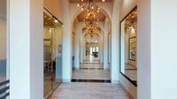 This thoughtfully designed single-floor space makes an immediate impression with its grand, arched hallway that serves as the architectural centerpiece. A series of graceful archways lines the corridor, creating a rhythmic, tunnel-like perspective that draws the eye toward a bright, open common area at the far end. Overhead, a procession of dramatic Sputnik-style chandeliers with warm Edison bulbs cast an inviting, ambient glow throughout the passage, adding a contemporary edge to the classical arch motif. The flooring features large-format light-toned tiles accented with darker inlay strips, lending the space a refined, polished character. To one side, floor-to-ceiling glass walls enclose office or meeting areas, maintaining visual openness while providing functional separation. Signage is visible through the glass, suggesting a professional or commercial use for the space. A large mirror on the opposite wall enhances the sense of depth and reflects the warm lighting beautifully. Natural light filters in from the far end of the corridor, brightening the space and hinting at additional open areas beyond. The overall ambiance is sophisticated and welcoming, balancing elegance with modern functionality.