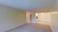 This single-floor residence offers a straightforward and functional layout designed for comfortable everyday living. The main living area is generously proportioned, featuring wall-to-wall carpeting in a neutral tone that adds warmth underfoot and complements the soft, light-colored walls throughout. The textured ceiling adds a classic touch to the space, contributing to its understated character. A wall-mounted heating unit is conveniently positioned in the living area, ensuring practical climate control. The open passage leading into the adjacent kitchen creates a natural flow between the living and culinary spaces. The kitchen, visible through the doorway, is well-lit and features white cabinetry and appliances, providing a clean and bright contrast to the warmer tones of the living area. The overall ambiance of the space is calm and neutral, offering a blank canvas well-suited for a variety of interior styles and furnishing arrangements. The single-floor layout ensures ease of movement throughout, making the space accessible and cohesive from room to room.