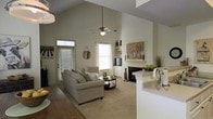 This two-story home makes an immediate impression with its dramatic vaulted ceilings in the main living area, creating a sense of openness and grandeur. The living room is a welcoming centerpiece, anchored by a classic fireplace with a light-toned surround and flanked by built-in shelving — ideal for displaying décor and media equipment. Large windows dressed with white blinds and sheer curtains invite generous natural light, enhancing the home's airy, neutral color palette of soft greens and warm whites. A ceiling fan adds both style and function to the soaring ceiling above. The open layout connects the living space to adjacent areas, with a partial dividing wall providing subtle separation while maintaining visual flow throughout. The flooring transitions from rich hardwood near the entryway to plush carpet in the main living zone, offering comfort underfoot. The overall ambiance is calm and inviting, blending casual elegance with practical design. With two floors, this home provides ample space for both everyday living and private retreats, making it a versatile and well-appointed residence suited to a variety of lifestyles.