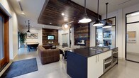 This single-floor space masterfully combines industrial and rustic design elements to create a warm yet contemporary environment. The open-concept layout seamlessly connects the kitchen, lounge, and dining areas, encouraging an easy flow throughout the space.

The kitchen is a standout feature, centered around a bold navy and white island with dark countertops and a professional-grade gas cooktop. Industrial-style pendant lights hang overhead, complementing the polished concrete floors that run throughout the space.

The lounge area is anchored by a rich brown leather sectional sofa and a striking reclaimed wood accent wall that frames a sleek, built-in gas fireplace. The same reclaimed wood material is carried up to the ceiling, forming a dramatic overhead feature that adds depth and warmth to the space.

Frosted glass partition doors provide a degree of privacy to adjacent areas while maintaining an open feel. A dining area with ample seating is positioned nearby, ideal for group gatherings. Recessed lighting combined with pendant fixtures ensures the space is well-lit throughout.

The overall ambiance is both inviting and sophisticated, striking a careful balance between rugged, natural textures and clean, modern finishes — making it well-suited for both casual use and professional or communal settings.