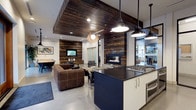 This single-floor space masterfully combines industrial and rustic design elements to create a warm yet contemporary environment. The open-concept layout seamlessly connects the kitchen, lounge, and dining areas, encouraging an easy flow throughout the space.

The kitchen is a standout feature, centered around a bold navy and white island with dark countertops and a professional-grade gas cooktop. Industrial-style pendant lights hang overhead, complementing the polished concrete floors that run throughout the space.

The lounge area is anchored by a rich brown leather sectional sofa and a striking reclaimed wood accent wall that frames a sleek, built-in gas fireplace. The same reclaimed wood material is carried up to the ceiling, forming a dramatic overhead feature that adds depth and warmth to the space.

Frosted glass partition doors provide a degree of privacy to adjacent areas while maintaining an open feel. A dining area with ample seating is positioned nearby, ideal for group gatherings. Recessed lighting combined with pendant fixtures ensures the space is well-lit throughout.

The overall ambiance is both inviting and sophisticated, striking a careful balance between rugged, natural textures and clean, modern finishes — making it well-suited for both casual use and professional or communal settings.
