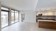 This contemporary single-floor space showcases a thoughtfully designed open-concept layout that seamlessly connects the living area and kitchen. Light hardwood flooring runs throughout, lending warmth and continuity to the space. Floor-to-ceiling sliding glass doors flood the living area with abundant natural light while offering direct access to an outdoor balcony with urban views, creating a strong connection between interior and exterior living.

The kitchen is a standout feature, boasting rich wood-grain cabinetry paired with a light-toned countertop and a striking mosaic tile backsplash in cool blue and grey tones. A kitchen island provides additional prep and seating space, while pendant lighting and track lighting above ensure the area is both functional and visually appealing. Stainless steel appliances, including a built-in microwave, complete the modern culinary setup.

White upper cabinetry adds brightness to the kitchen, complementing the clean white walls found throughout the space. A doorway leads to an additional room, suggesting a flexible layout suited for various living arrangements. The overall ambiance is clean, bright, and modern, reflecting a refined urban aesthetic ideal for contemporary living.