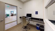 This single-floor business center is designed to provide a functional and professional workspace within a larger facility. The space is organized around two primary workstation areas, each equipped with a wall-mounted dark granite countertop surface, a computer, and a task chair, offering focused individual workspaces. A pass-through window connects one workstation alcove to an adjacent area, enhancing visibility and accessibility between zones.

A dedicated secondary room houses a multifunction printer and copier atop a matching dark countertop, keeping office equipment conveniently centralized. The walls throughout are finished in crisp white and soft blue-gray tones, creating a calm and professional atmosphere. Flooring transitions between neutral carpet in the work areas and light hardwood-style flooring in connecting corridors.

Glass-paneled doors connect the business center to a well-lit, furnished common lounge beyond, providing a sense of openness while maintaining separation between spaces. Colorful artwork adorns the walls, adding a welcoming touch to the otherwise utilitarian environment. Recessed and strip lighting ensures consistent illumination throughout, making the space practical for productivity at any time of day.