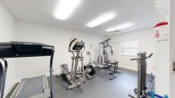 The Bell Oaks Fitness Zone is a well-appointed community fitness facility located in Montgomery, Alabama. The space is designed with functionality in mind, featuring clean white walls, durable dark rubber flooring, and bright overhead fluorescent lighting that ensures excellent visibility throughout workouts. A window with blinds allows natural light to supplement the artificial lighting, contributing to an open and energizing atmosphere.

The fitness area is thoughtfully equipped with a range of exercise machines to accommodate diverse workout routines. Cardio equipment includes a treadmill and an elliptical trainer, while strength training is supported by a multi-function cable and weight machine with an adjustable bench. Additional accessories such as exercise mats, resistance bands, stability balls, and a pull-up and dip station round out the available equipment.

A notable architectural detail is the vaulted ceiling in a connected area, featuring exposed white-painted wood beams, a skylight that invites natural light, and elegant hanging light fixtures — adding a touch of character to the overall space. The facility also includes a utility area accessible through an interior door, providing added convenience. Overall, the Bell Oaks Fitness Zone offers a clean, practical, and comfortable environment for residents to maintain an active and healthy lifestyle.