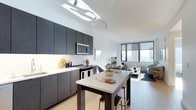 This single-floor space showcases a thoughtfully designed open-plan layout that seamlessly connects the kitchen, dining, and living areas into one cohesive environment. The contemporary kitchen is outfitted with dark, flat-front cabinetry that extends to the ceiling, providing ample storage while creating a bold visual contrast against the crisp white walls and light countertops. A central kitchen island with bar seating serves as both a functional workspace and an informal dining area, ideal for everyday meals or casual entertaining. Stainless steel appliances, including a built-in microwave and electric range, add a polished, professional touch. Track lighting overhead ensures the kitchen is well-lit throughout. The adjoining living area benefits from an abundance of natural light streaming through large floor-to-ceiling windows, which frame sweeping urban skyline views. Light hardwood-style flooring runs throughout, enhancing the sense of openness and warmth. The living space comfortably accommodates a seating arrangement, completing a layout that balances modern aesthetics with practical, day-to-day functionality. The overall ambiance is clean, bright, and metropolitan.