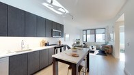 This single-floor space showcases a thoughtfully designed open-plan layout that seamlessly connects the kitchen, dining, and living areas into one cohesive environment. The contemporary kitchen is outfitted with dark, flat-front cabinetry that extends to the ceiling, providing ample storage while creating a bold visual contrast against the crisp white walls and light countertops. A central kitchen island with bar seating serves as both a functional workspace and an informal dining area, ideal for everyday meals or casual entertaining. Stainless steel appliances, including a built-in microwave and electric range, add a polished, professional touch. Track lighting overhead ensures the kitchen is well-lit throughout. The adjoining living area benefits from an abundance of natural light streaming through large floor-to-ceiling windows, which frame sweeping urban skyline views. Light hardwood-style flooring runs throughout, enhancing the sense of openness and warmth. The living space comfortably accommodates a seating arrangement, completing a layout that balances modern aesthetics with practical, day-to-day functionality. The overall ambiance is clean, bright, and metropolitan.