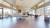 Located atop a building at 575 12th St S in Arlington, VA, this rooftop lounge offers a generously sized, single-floor entertainment space designed with a sophisticated modern aesthetic. The open-concept layout is anchored by rich hardwood-style flooring, soaring ceilings, and an abundance of natural light streaming through floor-to-ceiling glass walls and sliding doors that connect seamlessly to an outdoor terrace with sweeping views of the surrounding skyline.

The main lounge area features a striking arrangement of high-top bar tables with acrylic seating, plush lounge sofas, and a centerpiece billiards table beneath an elegant statement chandelier. A warm wood-paneled accent wall with an integrated large-screen display serves as a focal point, complemented by vertical wood slat detailing that adds architectural depth.

A dedicated kitchen and bar area is outfitted with a long counter lined with bentwood barstools, pendant globe lighting, and full appliance provisions, making it well-suited for catered events and social gatherings. A more intimate lounge nook, framed by dramatic floor-length red drapery, provides a private retreat within the larger space. Additional seating alcoves near the glass exterior walls offer direct access to the outdoor terrace, blurring the line between indoor comfort and open-air enjoyment.