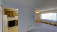 This single-floor space offers a straightforward and functional layout designed for comfortable everyday living. The living area is finished with wall-to-wall carpeting that adds warmth underfoot, and is illuminated by a ceiling fan with light fixture that provides both airflow and ambient lighting. A large window allows natural light to filter into the room, brightening the space and creating an open, airy feel. A baseboard heater runs along the wall, ensuring efficient climate control throughout the colder months. The textured ceiling adds a classic touch consistent throughout the space. Separated by a partial wall, the kitchen is distinct in its design, featuring vinyl tile flooring, warm-toned wood cabinetry, and black appliances including a dishwasher. The kitchen also includes a sink positioned beneath upper cabinets, making the most of the available space. The partial wall dividing the two areas provides a sense of definition between the living and cooking zones while maintaining an open connection between them. Overall, the space presents a practical and livable environment with a cohesive, understated aesthetic.
