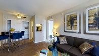 This single-floor residence offers a cohesive and thoughtfully designed living environment that balances comfort with modern style. The living area serves as the heart of the space, featuring rich hardwood flooring that extends throughout the main areas, creating a warm and inviting atmosphere. Smooth, light-toned walls provide a neutral backdrop that enhances the sense of openness, while a ceiling fan adds both function and a finishing touch to the room.

A built-in niche in the hallway wall adds an architectural detail that is both decorative and practical, offering a dedicated display space. The layout flows naturally between the living area and adjoining spaces, with clean sight lines connecting the main living room to what appears to be a kitchen and additional rooms beyond. The kitchen, visible in the background, features cabinetry and ample counter space.

The living room accommodates comfortable seating and is styled to highlight the space's versatility. Neutral tones are complemented by carefully chosen accent pieces, reinforcing the modern yet approachable character of the residence. Overall, the space presents a well-maintained, move-in-ready environment with a clean design sensibility and functional layout suited for everyday living.