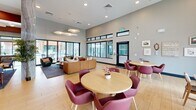 Located at 6856 West Maryland Avenue in Glendale, Arizona, this single-floor community clubhouse offers a generously sized, open-plan layout designed to accommodate a variety of social and recreational activities. The space is defined by its bold, contemporary design palette — warm wood-toned flooring flows throughout, while striking red-painted ceiling coffers and teal accent walls create a vibrant, energetic atmosphere.

The main gathering area features multiple seating arrangements, including round dining tables with deep burgundy chairs, comfortable lounge sofas, and café-style bistro tables positioned near floor-to-ceiling windows that invite abundant natural light and outdoor views of the surrounding community. A built-in banquette with gold upholstered paneling lines one wall, accompanied by a gallery of framed botanical artwork illuminated by wall-mounted picture lights.

A fully equipped kitchen anchors one end of the space, outfitted with stainless steel appliances, warm wood cabinetry, and a central island with bar seating beneath modern pendant lighting — ideal for resident gatherings and events.

A dedicated game room zone features a full-size pool table set atop a stylized area rug, surrounded by high-top seating and flanked by expansive sliding glass doors that open to outdoor views. Recessed and cove lighting throughout the space adds warmth and dimension, enhancing the overall inviting and social ambiance of this well-appointed clubhouse.