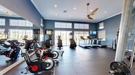 This fitness center offers a spacious, single-floor layout designed to accommodate a comprehensive range of workout activities. The interior is characterized by striking slate-blue walls, crisp white crown molding, and elevated ceilings that lend an open, airy feel to the space. Recessed lighting, combined with a large ceiling fan, ensures consistent illumination and airflow throughout. A bank of floor-to-ceiling windows and sliding glass doors lines one wall, flooding the room with natural light and providing outdoor views that enhance the workout atmosphere. The cardio zone features multiple treadmills, elliptical trainers, and stationary cycling bikes, each positioned to take advantage of the natural light. Wall-mounted flat-screen televisions are strategically placed for visibility from the cardio equipment. The strength training area is equally well-appointed, offering a cable machine system, a squat rack with weight plates, adjustable benches, a dumbbell rack, a punching bag, and a stability ball. Full-length mirrors line portions of the walls, visually expanding the space and supporting form-checking during exercise. Durable rubber flooring throughout provides both safety and functionality. A frosted glass partition elegantly divides zones within the room, adding a modern architectural detail while maintaining an open layout.