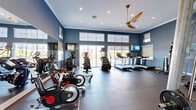 This fitness center offers a spacious, single-floor layout designed to accommodate a comprehensive range of workout activities. The interior is characterized by striking slate-blue walls, crisp white crown molding, and elevated ceilings that lend an open, airy feel to the space. Recessed lighting, combined with a large ceiling fan, ensures consistent illumination and airflow throughout. A bank of floor-to-ceiling windows and sliding glass doors lines one wall, flooding the room with natural light and providing outdoor views that enhance the workout atmosphere. The cardio zone features multiple treadmills, elliptical trainers, and stationary cycling bikes, each positioned to take advantage of the natural light. Wall-mounted flat-screen televisions are strategically placed for visibility from the cardio equipment. The strength training area is equally well-appointed, offering a cable machine system, a squat rack with weight plates, adjustable benches, a dumbbell rack, a punching bag, and a stability ball. Full-length mirrors line portions of the walls, visually expanding the space and supporting form-checking during exercise. Durable rubber flooring throughout provides both safety and functionality. A frosted glass partition elegantly divides zones within the room, adding a modern architectural detail while maintaining an open layout.