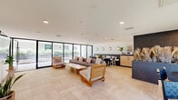 Located at 4971 S Centinela Ave in the Mar Vista neighborhood of Los Angeles, this single-floor communal space offers a thoughtfully designed open-plan layout encompassing a living room, dining area, and kitchenette. Large-format beige tile flooring flows seamlessly throughout, unifying the entire space with a clean, contemporary aesthetic.

The living area is a bright, welcoming focal point, furnished with a substantial sectional sofa, a low-profile wood-and-stone coffee table, and sculptural teak lounge chairs with plush cushions. Floor-to-ceiling black-framed sliding glass walls line one side of the room, flooding the interior with natural light and providing direct access to a landscaped outdoor courtyard, creating a seamless indoor-outdoor connection.

The dining zone features round black tables paired with woven wood chairs, set against a dramatic dark accent wall adorned with decorative dried pampas grass. Large windows on the adjacent wall offer views of surrounding greenery, further enhancing the sense of light and openness.

The kitchenette is neatly appointed with light wood cabinetry, dark stone countertops, and essential appliances, complemented by decorative wall sconces and potted fiddle-leaf fig plants. Recessed ceiling lighting throughout ensures a warm, ambient atmosphere. The overall design strikes a refined balance between modern minimalism and natural warmth.