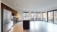 This single-floor unit is designed with a contemporary urban aesthetic, offering an open-concept layout that seamlessly integrates the kitchen, dining, and living areas. The expansive living space is finished with light hardwood-style flooring and benefits from an impressive wall of floor-to-ceiling windows along one side, flooding the interior with natural light and providing views of the surrounding cityscape. A sliding glass door leads to a private balcony, further connecting the interior to its urban surroundings.

The kitchen is well-appointed with rich dark-wood cabinetry, granite countertops, a penny-tile backsplash, and a full suite of stainless steel appliances including a gas range, microwave, dishwasher, and refrigerator. A large central island with matching granite provides additional workspace and storage.

The two bedrooms are carpeted and each features a large floor-to-ceiling window offering city views. The bathrooms are finished with granite-topped vanities and tile flooring. The primary bathroom includes a glass-enclosed walk-in shower, while the secondary bathroom features a tiled tub-shower combination. Track lighting throughout the unit provides consistent, adjustable illumination across all main living areas.