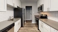 This well-appointed single-floor residence showcases a clean, contemporary aesthetic with consistent finishes carried throughout the space. The heart of the home is a generously proportioned galley-style kitchen, featuring floor-to-ceiling white shaker cabinetry with brushed nickel hardware, gray quartz countertops, and a classic white subway tile backsplash. A full suite of stainless steel appliances — including a refrigerator, electric range, over-the-range microwave, and dishwasher — provides a highly functional cooking environment. Wood-look luxury vinyl plank flooring runs throughout the kitchen and flows seamlessly into the adjoining dining area, which is illuminated by a chandelier and benefits from natural light through a large window overlooking greenery. The open living areas are finished with neutral gray walls, crisp white trim, and durable LVP flooring, while wall-mounted mini-split units provide efficient climate control. The bedroom offers plush carpeting, natural light from a window with outdoor views, and its own mini-split unit. The bathroom is outfitted with white subway tile surrounding a tub-shower combo, complemented by matching LVP flooring. The overall ambiance is modern, bright, and move-in ready.