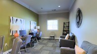 This single-floor space is designed to balance functionality and comfort in a thoughtfully arranged layout. The main room serves a dual purpose, seamlessly integrating a dedicated work area with a relaxed lounge setting. The workstation side features a spacious desk accommodating multiple monitors, paired with stylish grey task chairs, making it well-suited for focused work or collaborative use. A sleek black ladder-style shelving unit stands nearby, offering both storage and a decorative element. On the lounge side, a plush grey sofa with accent pillows invites relaxation, complemented by additional seating. A single window allows natural light to filter in, illuminating the room's neutral taupe and olive green accent walls, which lend a calm, grounded ambiance. Crown molding adds a refined architectural touch along the ceiling perimeter. Patterned carpet tiles cover the floor, adding texture and visual interest underfoot. Recessed lighting ensures the space remains bright and welcoming regardless of the time of day. Overall, the room presents a clean, modern aesthetic that supports a range of uses in a comfortable and organized environment.