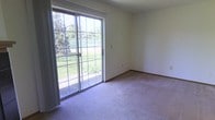 This two-story property offers a straightforward and functional layout across its two floors. The interior is finished with neutral-toned carpeting throughout, providing a soft, understated foundation that suits a variety of furnishing styles. The walls are painted in a light, off-white tone, contributing to a clean and bright atmosphere.

A standout feature of the space is the large sliding glass door, framed with a grid-pane design, which serves as both a source of abundant natural light and a direct access point to the outdoors. Through the glass, lush greenery, mature trees, and an open lawn area are clearly visible, creating a seamless connection between the interior and the surrounding landscape. The door appears to open onto a patio or ground-level exterior surface, making outdoor access convenient.

The room featuring the sliding door is generously proportioned, with enough space to accommodate a variety of furniture arrangements. The baseboards are finished in a contrasting darker tone against the light walls, adding a subtle architectural detail. Overall, the space presents a calm, neutral aesthetic with practical features and pleasant outdoor views.