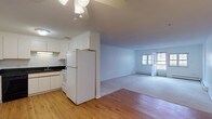 This single-floor, 2-bedroom, 1-bathroom residence offers a practical and well-organized layout designed for comfortable living. The kitchen is appointed with white cabinetry featuring gold-tone trim accents, dark granite-style countertops, a double sink, and white appliances including a refrigerator and range, complemented by a black dishwasher. Durable hardwood flooring runs through the kitchen and dining area, while a ceiling fan with light fixture adds functionality overhead. The kitchen opens into a spacious living area carpeted in neutral tones, which benefits from natural light streaming through windows and a glass door that leads to an outdoor balcony. Baseboard heating is present throughout the home. Both bedrooms are generously sized, each featuring plush wall-to-wall carpeting, large windows that invite natural light, and dedicated closets with built-in rods for ample storage. The full bathroom features a classic black-and-white octagonal tile floor, a white vanity, a toilet, and a tub-shower combination with a curtain rod. The overall color palette throughout the home is clean and neutral, with white walls creating a bright, airy ambiance that offers a versatile canvas for personalization.