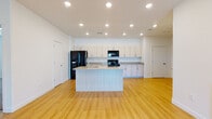 This modern single-floor residence offers a thoughtfully designed open-concept layout that seamlessly connects the kitchen and living areas. The kitchen serves as a central hub, anchored by a large island with a granite countertop and an integrated sink, providing ample workspace and casual seating. Light wood-tone cabinetry lines the walls, complemented by granite countertops, black appliances including a refrigerator, electric range, and built-in microwave. Recessed lighting throughout the kitchen and living areas creates a bright, welcoming atmosphere, while light hardwood flooring adds warmth and continuity across the open space. A ceiling fan in the living area enhances comfort, and sliding glass doors offer access to an outdoor balcony. The bedrooms are finished with plush neutral carpeting and feature windows with blinds that allow for natural light and privacy. Each bedroom includes closet storage. The bathrooms are well-appointed with light wood vanities, granite countertops, large wall mirrors, and hardwood flooring, maintaining a consistent and cohesive aesthetic throughout the residence. Clean white walls and a neutral color palette give the entire space a fresh, contemporary feel.