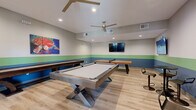 This dedicated game room is a well-appointed recreational space designed for social entertainment and leisure. The room features light hardwood-style flooring throughout, complemented by a distinctive tri-color horizontal stripe along the walls — combining soft gray, green, and blue tones — that gives the space a modern, energetic character. Recessed lighting and dual ceiling fans ensure the room remains well-lit and comfortable during extended use.

At the center of the room sits a contemporary pool table with a sleek metal base and a clean white felt surface, serving as the focal point of the space. A wooden shuffleboard table runs along one wall, while a classic ping pong table occupies a dedicated corner, providing a variety of game options for guests. Multiple wall-mounted flat-screen televisions are positioned around the room, allowing guests to watch sports or other programming while playing.

High-top tables with bar stools are arranged along one wall, offering comfortable seating for spectators or those taking a break between games. A cue rack and additional accessories are neatly stored nearby. Vibrant, sports-themed artwork adds personality to the walls. Glass-panel doors connect the game room to an adjacent fitness area, enhancing the overall sense of an active, amenity-rich environment.