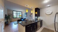This single-floor residence is thoughtfully designed with an open-concept layout that seamlessly connects the kitchen, dining, and living areas. The kitchen serves as a central hub, showcasing dark espresso cabinetry, dark granite countertops, a tile mosaic backsplash, and stainless steel appliances including a refrigerator, range, and built-in microwave. A peninsula with bar seating provides additional prep space and casual dining, illuminated by two striking yellow pendant lights that add a bold design accent. Light hardwood-style flooring flows throughout the main living areas, contributing to a clean, modern aesthetic.

The living area receives generous natural light through large windows overlooking an outdoor courtyard, creating an inviting and airy atmosphere. The bedroom features plush carpeting, a large window with blinds, and ample room for a full furniture arrangement. The bathroom maintains design consistency with matching dark cabinetry, granite countertops, a single vanity with mirror, and a tub-shower combination with tile surround. Throughout the unit, neutral wall tones provide a versatile backdrop that complements the contemporary finishes.