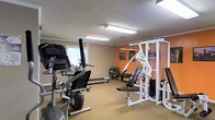 This fitness center offers a practical and well-organized workout environment spread across a single floor. The space is designed to accommodate a range of exercise needs, featuring a mix of cardio and strength training equipment. Cardio options include an elliptical machine and a treadmill, positioned to take advantage of natural light from a window along one wall. A large, multi-station cable and weight machine anchors the strength training area, complete with an adjustable bench and a substantial weight stack, making it suitable for a variety of resistance exercises. The flooring throughout is a durable, cushioned carpet in a neutral tan tone, providing comfort underfoot during workouts. Walls are painted in a combination of warm beige and an energizing orange accent, contributing to a motivating atmosphere. Overhead, fluorescent lighting ensures the space is evenly and brightly illuminated. A textured ceiling and decorative wall art add subtle character to the room. A small potted plant near the corner introduces a touch of greenery, softening the utilitarian nature of the space. Overall, the fitness center strikes a balance between functionality and a welcoming ambiance.
