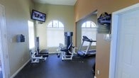This single-floor fitness center offers a functional and well-organized workout space designed to accommodate a variety of exercise needs. The room is characterized by its warm, neutral-toned walls and durable rubber flooring, which provides both safety and comfort during workouts. Arched windows allow natural light to filter into the space, creating a bright and inviting atmosphere while offering views to the outside. A wall-mounted flat-screen television is positioned for easy viewing during cardio sessions, adding an entertainment element to the workout experience. The space is equipped with a treadmill for cardiovascular training alongside multiple strength training machines, including what appears to be a leg press and cable weight station, catering to both upper and lower body workouts. A clearly displayed "Fitness Center Rules" sign is posted near the entrance, ensuring users are informed of facility guidelines. The overall layout is efficiently arranged to maximize the available floor space, making the most of the room's footprint while maintaining clear pathways between equipment. The space presents a clean, practical environment suited for everyday fitness use.