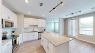 This single-story residence in Pensacola, Florida offers a thoughtfully designed open-concept layout with contemporary finishes throughout. The kitchen serves as a central highlight, showcasing white shaker-style cabinetry with brushed nickel hardware, speckled granite countertops, a subway tile backsplash, and a full suite of stainless steel appliances including a refrigerator, range, microwave, and dishwasher. A center island provides additional prep and seating space, while a deep undermount stainless steel sink with a professional-style faucet adds a functional touch. Track lighting and recessed fixtures illuminate the space effectively. The kitchen flows seamlessly into a bright, open living area featuring light wood-style plank flooring, neutral walls, and a ceiling fan. Natural light enters through large windows that frame views of the surrounding greenery. The bedrooms offer plush carpeting, ceiling fans, and windows with privacy blinds, creating comfortable retreats. The bathrooms are finished with granite-topped dual vanities, white cabinetry, large mirrors, and frameless glass-enclosed showers adorned with elegant marble-style tile surround. Wood-look flooring continues into the bathrooms, tying together the cohesive, modern aesthetic found throughout this well-appointed residence.