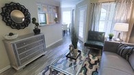 This single-story space showcases a thoughtfully designed open-concept layout that connects the living and dining areas with a natural, cohesive flow. The living area features rich hardwood-style flooring that extends throughout, anchored by a geometric-patterned area rug in cool blue and grey tones. A glass-topped coffee table with a metallic frame serves as a central focal point, complemented by a tufted leather accent chair in deep teal. A mirrored dresser along one wall adds a reflective, contemporary touch to the space.

Natural light fills the interior through multiple French doors dressed with patterned curtains, creating an airy and welcoming ambiance. Crown molding detail along the upper walls adds an element of architectural refinement. A pass-through window connects the living area to a raised breakfast bar, offering a glimpse into the dining space beyond, where bar-height seating and additional windows contribute to the bright, open feel. Decorative greenery and sculptural accents are thoughtfully placed throughout, enhancing the modern yet approachable character of the space.