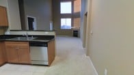 This two-storey space showcases an impressive open-concept layout where the kitchen flows seamlessly into the living area. The kitchen is equipped with dark granite countertops, warm wood cabinetry, a stainless steel dishwasher, and a functional island. Tile flooring anchors the kitchen space, transitioning into plush carpeting throughout the living area.

The living area benefits from dramatic double-height ceilings, which create a grand sense of openness and volume. Large windows, including an oversized upper-level window, flood the space with natural light while offering views of the surrounding neighbourhood. A fireplace serves as a focal point in the living area, adding warmth and character to the room.

The warm taupe wall tones complement the neutral flooring and cabinetry, creating a cohesive and inviting atmosphere throughout. The vertical height of the space, accentuated by the two-storey design, contributes to an airy and expansive feel that makes the interior feel both functional and welcoming.