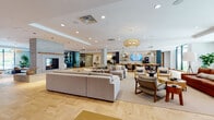 This expansive single-floor amenity lobby is thoughtfully designed to serve as a dynamic communal hub, blending comfort, function, and contemporary style. The space is unified by striking herringbone-patterned hardwood flooring that flows seamlessly throughout, anchoring the various zones with warmth and visual continuity.

The main lounge area is generously appointed with oversized sectional sofas, accent chairs, and layered area rugs, all centered around a sleek stone fireplace that serves as a focal point. Woven rattan pendant lights hang overhead, adding a natural, organic touch to the modern interior. Large floor-to-ceiling windows flood the space with abundant natural light and offer views of the surrounding greenery outdoors.

Adjacent to the lounge, a well-equipped kitchenette and bar area features open shelving, pendant lighting, and bar-height seating, making it ideal for casual gatherings. A glass-enclosed room with frosted sliding doors provides a private space suitable for meetings or focused work, outfitted with a dining-style table and task lighting.

A dedicated mailroom area includes an extensive bank of individual mailboxes set within a clean, white built-in surround, illuminated by additional woven pendant fixtures. Curated artwork, lush potted plants, and thoughtful décor details complete the polished, resort-inspired ambiance of this well-appointed communal space.