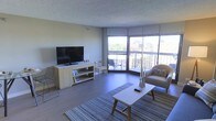 This single-floor space centers around a well-appointed living area designed to maximize natural light and openness. Large floor-to-ceiling windows wrap around one corner of the room, flooding the interior with daylight and framing views of the surrounding urban and green landscape. A sliding glass door leads directly to a private balcony, seamlessly connecting the indoor living area to the outdoors.

The living room features smooth, light-toned hardwood-style flooring that complements the neutral wall tones and textured ceiling. The space is laid out to accommodate comfortable seating, with a cushioned armchair and a streamlined coffee table anchoring the area over a woven area rug. A low-profile media console houses a flat-screen television, and additional seating options, including a wooden accent chair, add both function and style.

The overall ambiance is calm and contemporary, with a minimalist aesthetic supported by clean lines and a muted, earthy color palette. Natural light plays a central role in defining the character of this space, making it feel welcoming and expansive throughout the day.