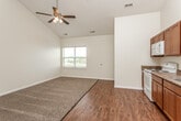 This single-floor space is defined by its striking vaulted ceilings, which lend an airy, open feel to the main living and kitchen area. The living space combines plush carpeting with rich hardwood-style flooring, creating a clear visual distinction between the lounge and kitchen zones. A ceiling fan with a light fixture is centrally positioned overhead, providing both airflow and ambient lighting. A window along one wall allows natural light to filter in, brightening the neutral, cream-toned walls throughout.

The kitchen runs along one wall in a galley-style configuration, featuring warm medium-toned oak cabinetry that extends both above and below the countertops. Stone-patterned laminate countertops provide ample workspace, complemented by a double stainless steel sink, a white electric range with overhead microwave, a dishwasher, and a full-size refrigerator. Recessed lighting adds additional illumination to the kitchen.

The bathroom is finished with matching oak cabinetry, a single vanity with a light stone-toned countertop, a wood-framed mirror, and tile flooring in a neutral beige tone. A washer and dryer area is also visible within the unit, adding practical convenience to the overall layout.