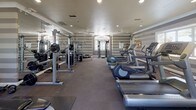This fitness center is a comprehensively outfitted exercise facility designed to support a full range of workout routines. The space is distinguished by its bold horizontal striped walls in alternating shades of gray and taupe, lending a polished, modern aesthetic throughout. Recessed lighting and ceiling fans are installed overhead, ensuring the space remains well-lit and comfortable during use.

Natural light filters in through multiple white-shuttered windows, offering views of the surrounding greenery outside. Full-length mirrors along one wall visually expand the space and allow for form monitoring during exercise.

The cardio section features multiple treadmills, elliptical trainers, and stationary bikes positioned on protective rubber flooring mats. The strength training area is well-appointed with cable and pulley machines, a multi-station weight machine, a free-weight rack stocked with dumbbells, and flat benches for a variety of exercises. Additional accessories including stability balls and resistance tools are available for supplemental training.

A wall-mounted television provides entertainment, while a bulletin board offers informational postings for users. The carpeted flooring throughout the main area, combined with protective mats under equipment, contributes to a clean and functional environment. The overall layout is spacious and organized, allowing multiple users to exercise simultaneously without congestion.