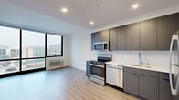 Located at 9 Homestead Place in Jersey City, NJ, this modern one-bedroom, one-bathroom residence offers a thoughtfully designed layout with contemporary finishes throughout. Light wood-tone flooring flows seamlessly across the open-plan kitchen and living area, creating a cohesive and inviting atmosphere. The kitchen is well-appointed with dark wood grain cabinetry extending floor-to-ceiling, stainless steel appliances including a range, over-the-range microwave, dishwasher, and refrigerator, complemented by a crisp white tile backsplash and light countertops. Floor-to-ceiling windows in the living area and bedroom flood the space with natural light while offering striking elevated views of the Jersey City skyline and surrounding urban landscape, with glimpses of the New York City skyline visible in the distance. The bedroom is a serene retreat, featuring expansive windows and clean white walls. The bathroom is finished with large-format tile flooring, a dark vanity with mirror cabinet, and a glass-enclosed walk-in shower with white subway tile. Additional conveniences include two closets and an in-unit laundry area, rounding out this efficient and modern urban residence.