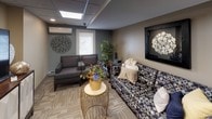 This community space is thoughtfully designed to serve as a versatile gathering and recreational hub, all on a single floor. The main lounge area sets a welcoming tone with comfortable seating, including a plush sofa and armchairs arranged around a decorative coffee table, complemented by a wall-mounted flat-screen TV atop a stylish media console. Neutral-toned carpeting, recessed lighting, and a drop ceiling with skylight panels create a bright, inviting ambiance throughout. A private meeting room, accessible through sleek dark-framed glass doors, offers an intimate setting with a round table, cushioned chairs, and tasteful wall art — ideal for small gatherings or quiet conversation. The kitchenette is fully equipped with dark cabinetry, granite-style countertops, a stainless steel refrigerator, microwave, coffee maker, and a double sink, making it convenient for hosting events or casual get-togethers. A split-unit air conditioner ensures year-round comfort in the lounge. Decorative accents, including wall art, potted plants, and curated décor pieces, add a polished, residential feel to the space. Together, these areas provide a functional and aesthetically cohesive environment for community members to gather, collaborate, and unwind.