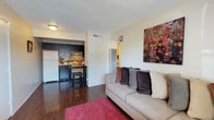 This single-floor unit offers an open-concept layout that seamlessly connects the living area and kitchen, creating a functional and inviting space. The living area features rich, dark hardwood flooring that extends throughout, complemented by neutral-toned walls that lend a clean and versatile backdrop. A comfortable sofa anchored by a vibrant red area rug defines the seating area, while bold wall art adds a pop of color and personality to the space.

The kitchen is efficiently designed with dark espresso cabinetry on both upper and lower sections, providing ample storage. A mosaic tile backsplash adds visual interest, and a white refrigerator anchors one end of the kitchen. A compact breakfast bar with bar stools serves as a casual dining option and a natural divider between the kitchen and living space.

A textured popcorn ceiling with flush-mount lighting provides overhead illumination, while a visible HVAC vent ensures climate control. A hallway visible in the background hints at additional rooms, possibly a bedroom and bathroom. Overall, the space balances practicality and style in a cozy, efficiently utilized layout.