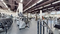 This comprehensive two-floor fitness facility is designed to accommodate a wide range of training disciplines across generously proportioned open floor plans. The industrial-style architecture is characterized by exposed ceilings with visible ductwork and structural beams, bright linear fluorescent lighting, and durable rubber flooring throughout — creating a functional and energetic workout environment.

The cardio zone features a robust lineup of stair climbers, ellipticals, and other aerobic machines arranged for easy access and flow. A dedicated cycling studio houses rows of stationary spin bikes facing wall-to-wall mirrors, offering an immersive group fitness experience. The strength training areas are extensive, with separate sections for selectorized machines, free weights with multiple benches and dumbbell racks, and a full power lifting zone equipped with squat racks, barbells, and plate-loaded stations.

A functional training room provides a clean, open mat space suited for agility drills, sled work, tire flips, and medicine ball training. Large windows positioned along the upper walls allow natural light to filter in, enhancing the bright and airy atmosphere. Mirrored walls throughout amplify the sense of space and support form monitoring. Staircase access connects the two floors seamlessly, completing a well-organized, full-service fitness destination.