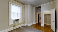 This single-floor space showcases a blend of classic and functional design elements throughout. The interior features warm hardwood flooring in the main living and corridor areas, complemented by neutral-toned walls and crisp white baseboards and crown molding that add a traditional architectural character to the space.

The main living area is bright and open, benefiting from a large window fitted with blinds that allows natural light to fill the room. A wall-unit air conditioner provides climate control, and the room transitions seamlessly between carpeted and hardwood floor sections, offering distinct zones within the space.

A compact powder room is accessible from the interior, outfitted with a wall-mounted sink and a sconce light fixture, providing a clean and practical layout. White paneled doors throughout the space contribute to a cohesive and classic aesthetic.

The corridor connects the various areas efficiently, with glimpses of a closet space and additional rooms beyond, suggesting a well-organized and functional floor plan. Overall, the space carries a neutral, understated ambiance with its warm tones, traditional trim work, and practical features — making it a versatile and livable environment suited to a variety of uses.