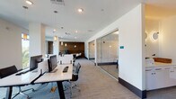 Located at 3110 Grand Ave in Pinellas Park, Florida, this contemporary single-floor office space is thoughtfully designed to support a variety of work styles. The open-plan layout features rows of sleek, white workstations equipped with monitors and ergonomic seating, providing a clean and efficient environment for collaborative or focused work.

Flanking the main workspace are floor-to-ceiling glass-walled private offices, offering visual transparency while maintaining acoustic separation. These enclosed offices are bright and minimally furnished, making them adaptable for individual use or small team meetings.

A standout lounge area incorporates high-back booth-style seating positioned against a richly textured wood-plank accent wall, creating an inviting space for informal discussions or focused work. Nearby, a separate seating nook features a wall-mounted flat-screen TV, a round coffee table, and soft armchairs arranged around a vibrant teal accent wall, ideal for presentations or casual gatherings.

Large windows throughout the space allow generous natural light to fill the interior while offering views of the surrounding Florida landscape. A dedicated service area with built-in cabinetry and office equipment adds practical functionality. Pendant lighting, patterned carpet tile flooring, and a neutral, modern color palette complete the polished, professional ambiance of this well-appointed office space.