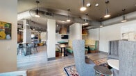 Located at 4090 Rio Vista Dr in Colorado Springs, CO, this single-floor community space offers a well-appointed and thoughtfully designed lounge and kitchen area. The open-concept layout is anchored by light hardwood-style flooring that flows throughout, complemented by an industrial-chic aesthetic defined by exposed ductwork, bold dark ceiling accents, and an array of stylish pendant lighting in varied shapes and finishes.

The living area is divided into distinct yet connected zones. A comfortable seating area features high-back wingback chairs arranged over vibrant patterned area rugs, creating an inviting spot for relaxation or conversation. Adjacent to this is a striking dark marble-tile fireplace wall, adding warmth and sophistication. A pool table serves as a central social focal point, surrounded by booth-style seating with green upholstered benches and mosaic tile backsplashes, ideal for group gatherings. Wall-mounted televisions are positioned throughout for added convenience.

The kitchen is a standout feature, showcasing bold navy-blue cabinetry with brushed gold hardware, crisp white quartz countertops, and a white geometric tile backsplash. A large center island provides ample workspace, while a generous window floods the area with natural light and offers views of an outdoor patio. The space combines functionality with striking visual design, making it equally suited for casual use or hosted events.