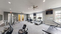 Located at 6334 Clock Tower Dr in Walbridge, Ohio, this well-equipped fitness center spans a single floor and offers a comprehensive workout environment. The main exercise room is generously proportioned, featuring neutral gray walls, patterned carpet tile flooring, and recessed lighting complemented by dual ceiling fans that maintain air circulation throughout the space. Large multi-pane windows line the walls, filling the room with natural light and providing views of the surrounding exterior. The cardio zone is outfitted with multiple treadmills, elliptical trainers, and a stationary bike, with wall-mounted televisions positioned for easy viewing during workouts. A dedicated strength training area features a full rack of dumbbells displayed on tiered stands, a multi-function cable machine, and a weight bench, all set on dark rubber flooring and backed by floor-to-ceiling mirrors that enhance the sense of space. A separate room houses a tanning bed, providing an additional amenity for users. Glass doors connect the fitness center to an adjacent leasing or community office area, reinforcing its role as part of a larger residential community. Workout schedules and informational boards are posted for member reference, rounding out this functional and well-maintained facility.