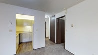 This single-floor residential unit offers a straightforward and functional layout designed for comfortable everyday living. The interior features a combination of hard surface flooring and carpet, providing a practical mix of textures across the living and transitional spaces.

The kitchen is situated in an alcove-style nook, outfitted with light-colored cabinetry, under-cabinet lighting that casts a warm glow over the countertops, a sink, and a full-size white refrigerator. The compact yet efficient design makes good use of the available space.

Adjacent to the kitchen, a central hallway connects the main living areas, anchored by dark wood-toned doors that add a contrasting warmth to the predominantly white walls. The hallway leads to what appears to be additional rooms or closets, including a set of dark panel closet doors that offer ample storage. The white walls and standard-height ceilings keep the interior feeling clean and neutral throughout.

The open transitional area between the kitchen and hallway features carpeting underfoot, creating a softer zone within the unit. Overall, the space presents a clean, move-in ready environment with a classic, no-frills aesthetic well-suited for practical residential use.