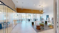 This thoughtfully designed single-floor workspace offers a generous open layout that seamlessly blends focused work areas with collaborative lounge zones. The space is anchored by a series of fully glazed, floor-to-ceiling glass-walled meeting rooms, providing transparency and a sense of openness while still offering private enclosed settings for team collaboration. Each glass-enclosed room is equipped with conference-style tables and ergonomic seating, bathed in natural light streaming in through expansive windows that frame lush outdoor views.

Warm oak wood paneling lines the upper walls throughout, adding a natural, grounding contrast to the crisp white ceilings and light hardwood flooring below. The open lounge areas feature a curated mix of colorful, modern furnishings — including rounded ottomans in muted teals, pinks, and greens — complemented by sculptural pendant lighting and contemporary accent tables.

Cozy built-in banquette nooks with white tulip-style tables provide semi-private spots for informal meetings or individual work. A comfortable lounge corner with a sectional sofa, vibrant patterned area rug, and floor-to-ceiling windows adds a residential warmth to the space. Curated artwork in bold primary colors adorns the walls, reinforcing the creative and energetic atmosphere. The overall ambiance is bright, airy, and professionally inviting.